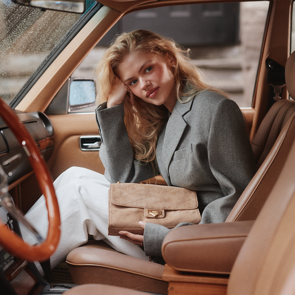 A woman sits in the front passenger seat of a vintage car with tan leather interiors, holding a taupe suede bag with a gold-tone turn-lock clasp.