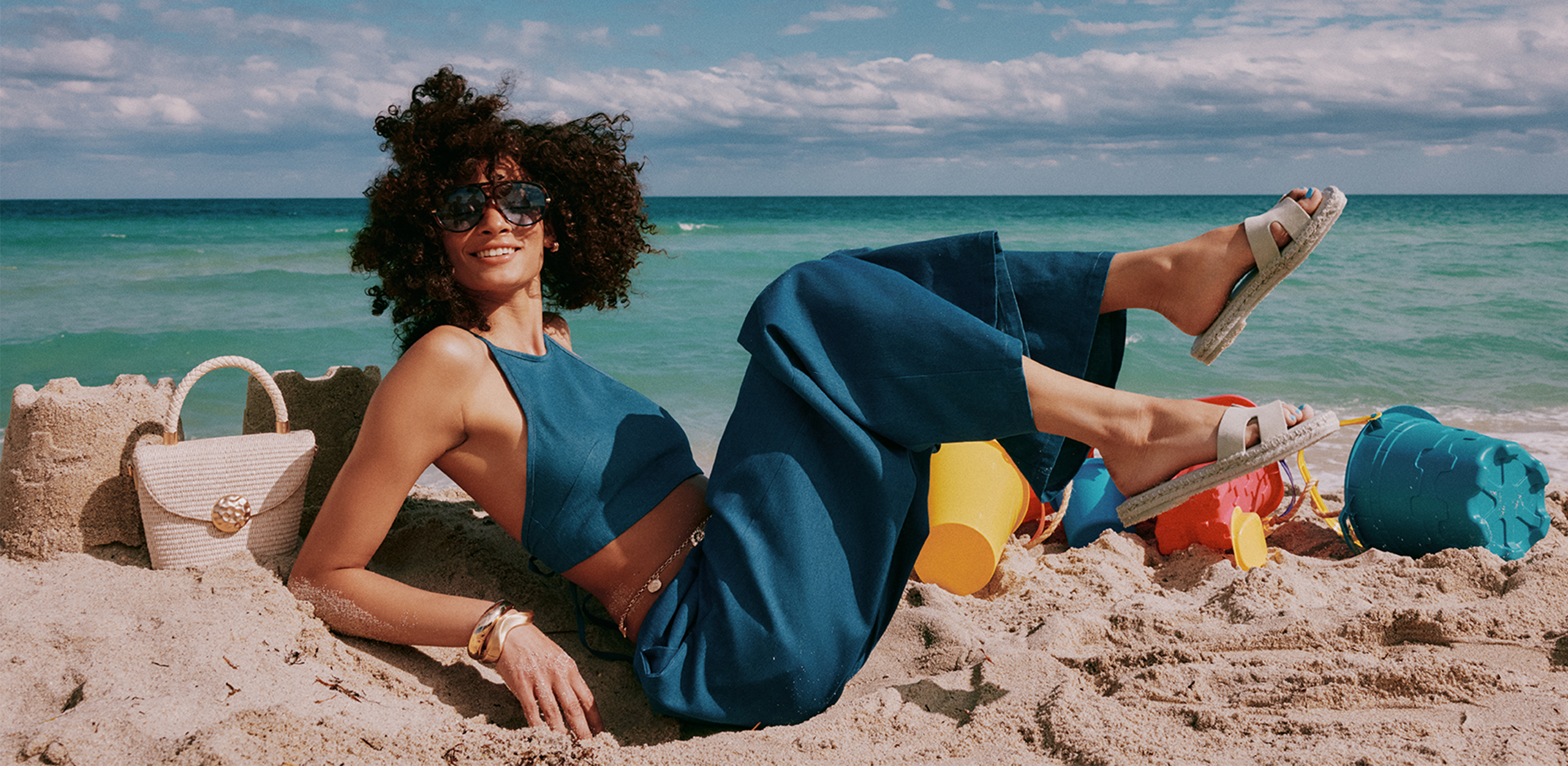 Woman in a blue outfit lying on the beach with a colorful bag and sandals.