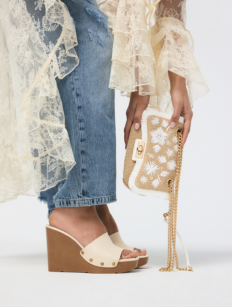 Person wearing beige wedge sandals and holding a gold handbag with lace detailing.