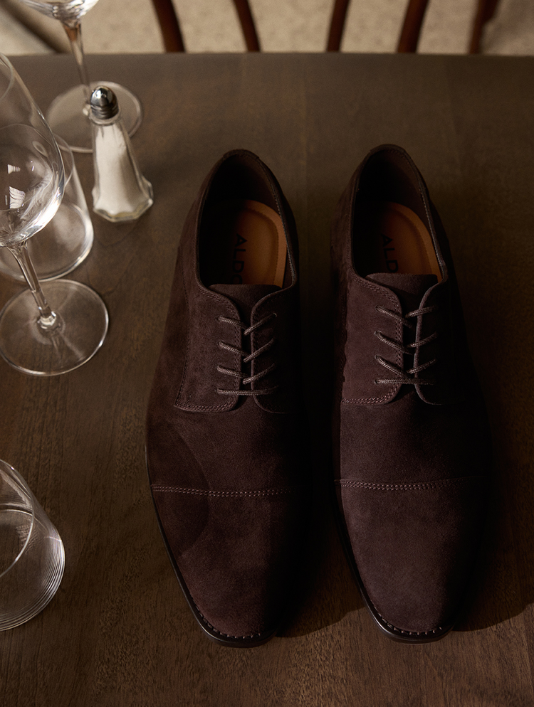 A pair of dark brown suede oxford shoes with matching laces, displayed on a dark wooden table with wine glasses and a salt shaker in the background.