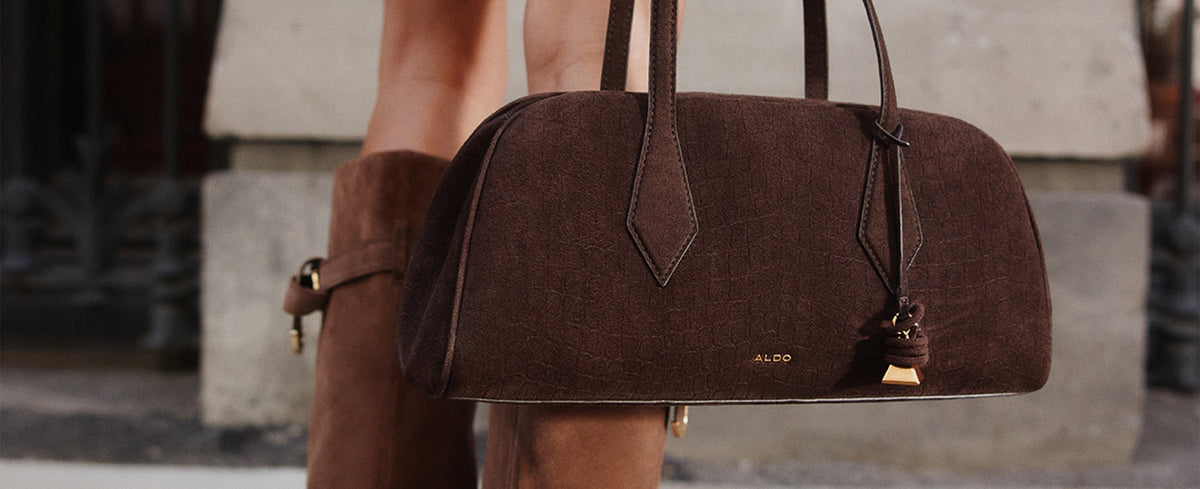 A dark brown suede duffle-style handbag with double shoulder straps, a small gold-tone logo plaque, and a hanging charm, carried by a model wearing matching brown suede boots against a blurred stone background.