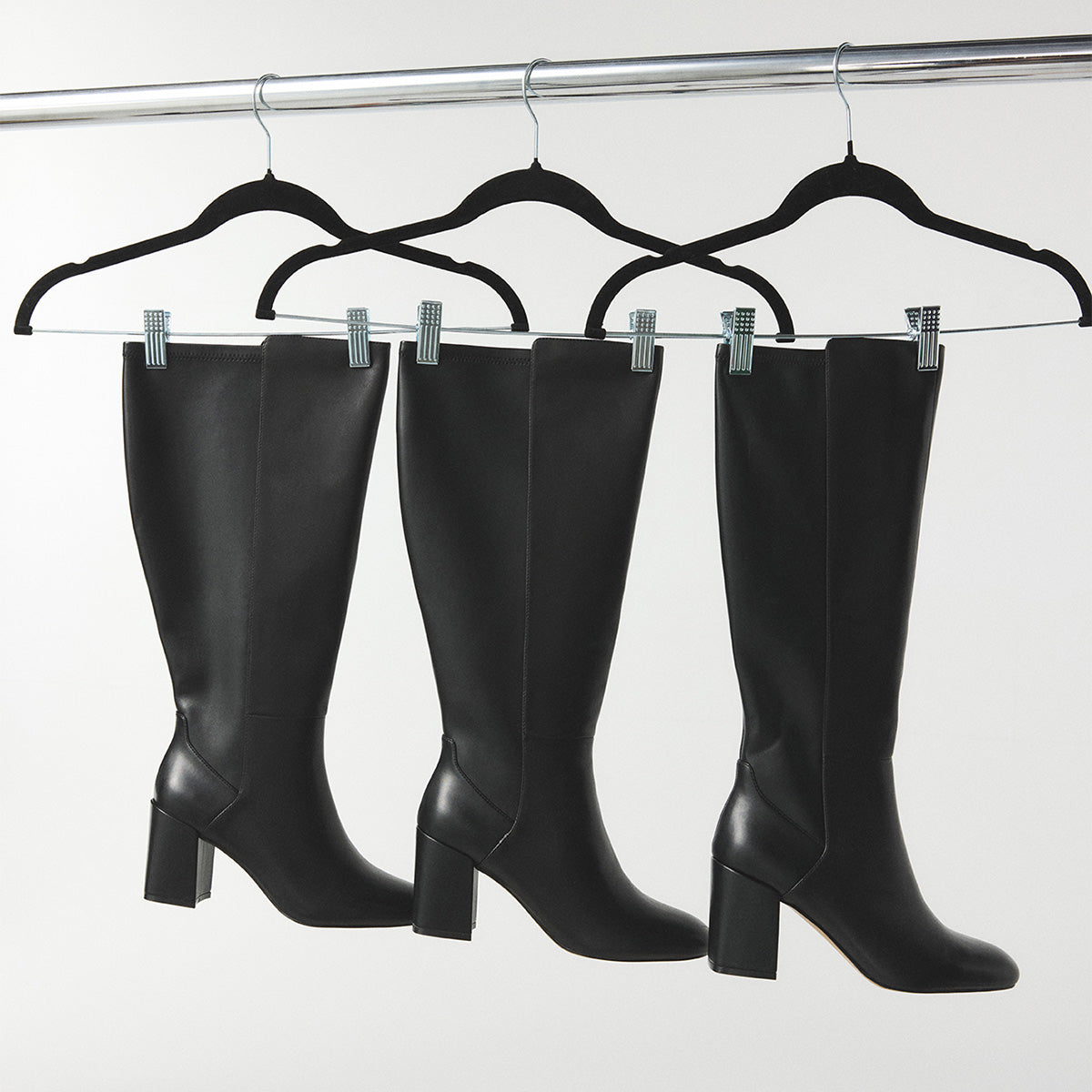 Three black knee-high leather boots with block heels hanging from black hangers on a silver clothing rack against a light gray background.