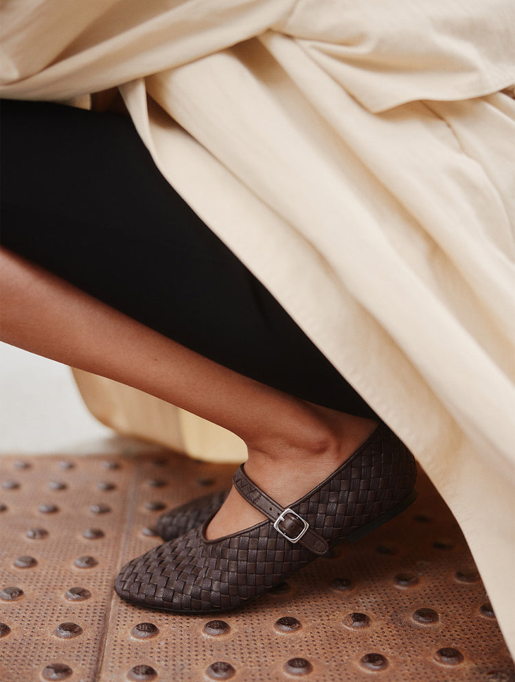 A close-up of a person wearing dark brown woven Mary Jane flats with a silver buckle, seated under draped cream fabric on a brown perforated surface.