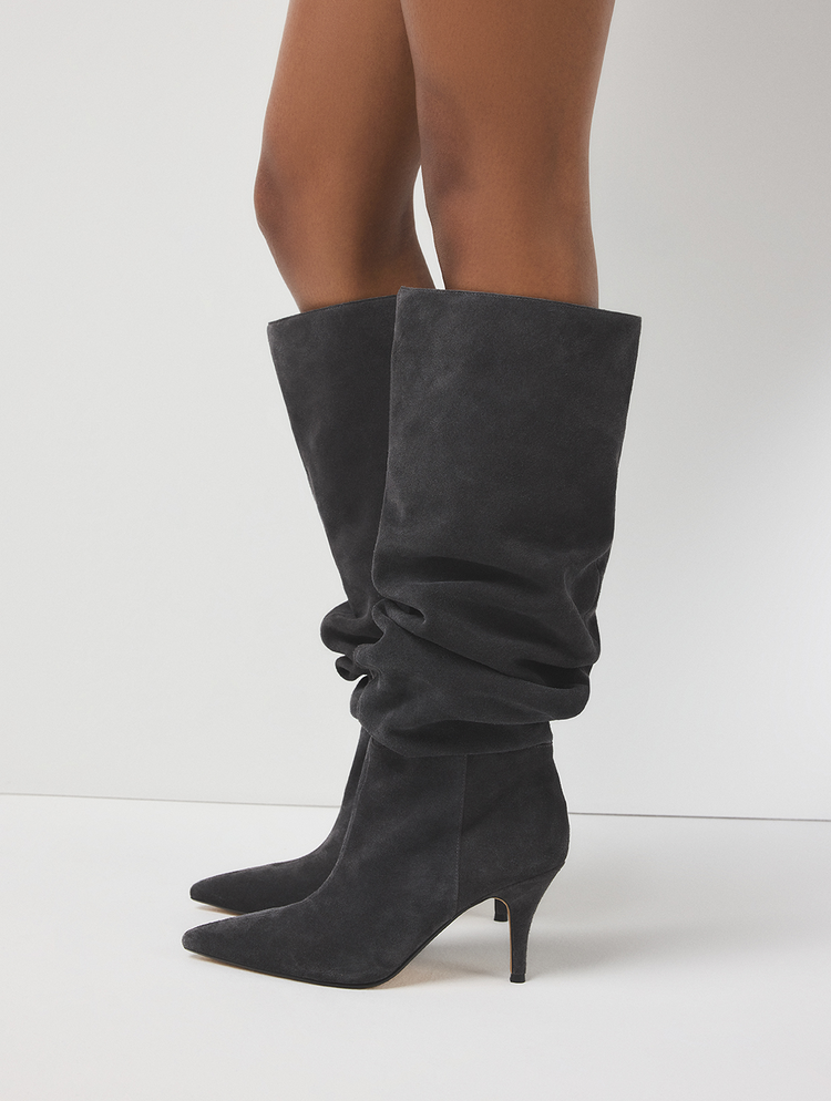 A pair of dark gray suede knee-high slouch boots with pointed toes and kitten heels, shown on a model's legs against a clean white studio background.