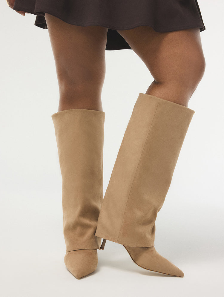 A person wearing tall knee-high pointed-toe heeled boots in a light tan suede-like finish, shown against a plain light gray studio background.